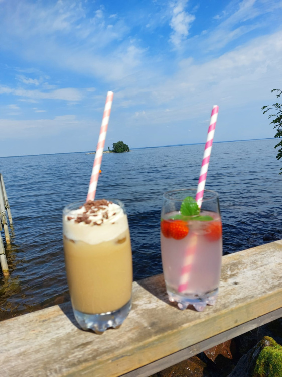 Kaksi kesäjuomaa puukaiteella. Taustalla näkyy järvimaisemaa.
